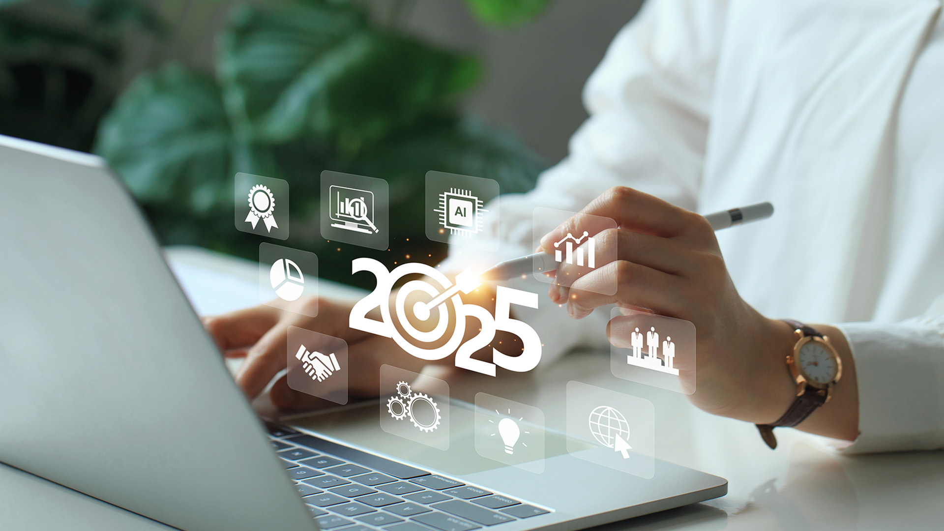 A person sitting at a laptop using a stylus to create a stylized 2025 surrounded by various business-related icons